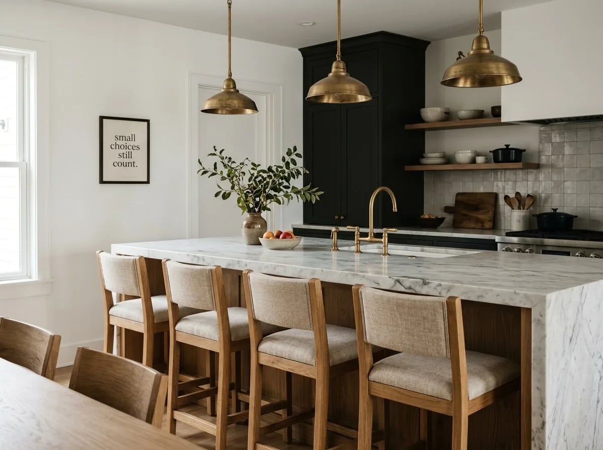A marble kitchen island with four upholstered wooden stools under brass pendant lights and a framed sign reading 'small choices still count'